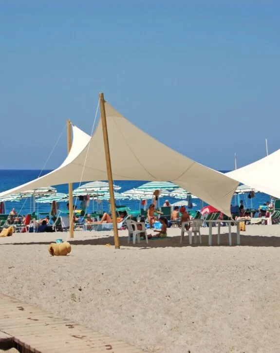 Spiaggia affollata con tende bianche e ombrelloni sopra sabbia chiara e mare blu sullo sfondo.