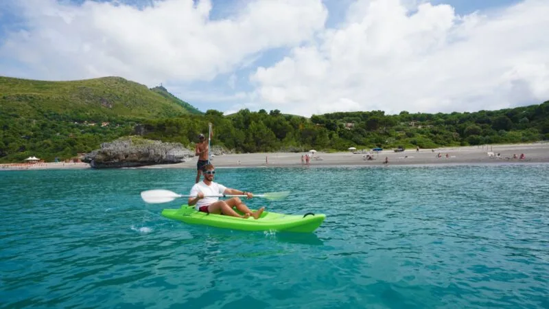 Due persone in kayak sulla spiaggia con fondale verde e colline sotto un cielo nuvoloso.