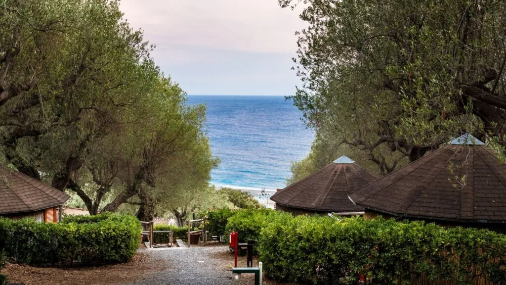 Vista di un sentiero con capanne di legno e alberi che guarda verso il mare.