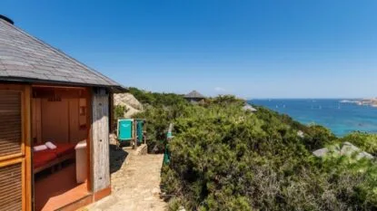 Struttura in legno con vista sul mare, circondata da vegetazione e barche a vela in lontananza sotto un cielo azzurro.