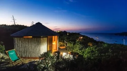 Baita con pareti in legno, illuminata dalla luce calda, affacciata su un mare sereno al crepuscolo.