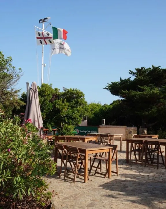 Area all'aperto con tavoli in legno, bandiere italiane e un'insegna del Touring Club Italiano, circondata da vegetazione.