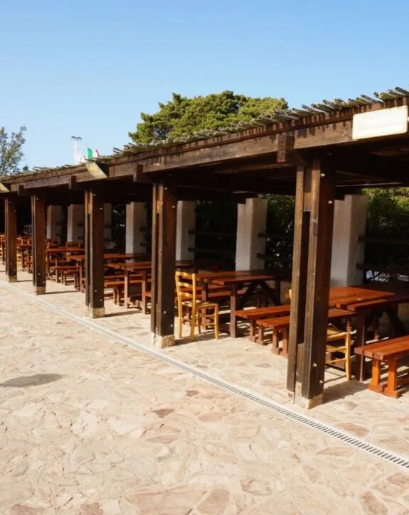 Area esterna di un ristorante con tavoli in legno, sedie e pergolato, sotto un cielo azzurro.