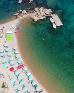Spiaggia con ombrelloni a strisce, kayak colorati e un pontile affacciato su acque cristalline. Giocatori di spiaggia visibili.