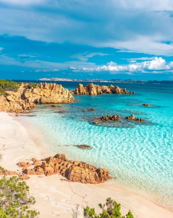 Spiaggia con sabbia bianca e acque turchesi, circondata da rocce e vegetazione, sotto un cielo sereno e nuvoloso.