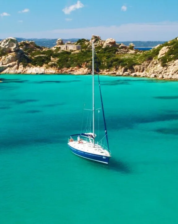Una barca a vela blu naviga in acque turchesi con rocce e vegetazione sullo sfondo. Cielo sereno e sole splendente.