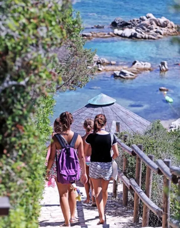 Quattro ragazze camminano lungo un sentiero verso una spiaggia con acque turchesi e rocce, sotto un cielo sereno.