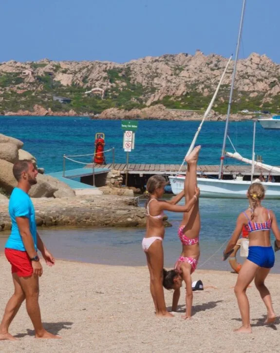 Bambini giocano sulla spiaggia; due stanno facendo una verticale mentre un adulto osserva e altre due ragazze si divertono.