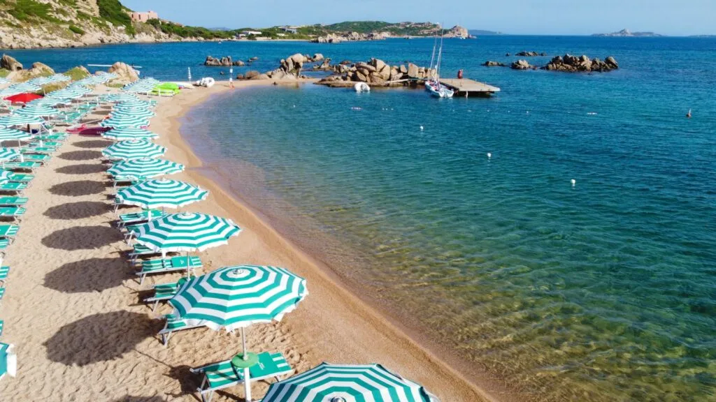 Spiaggia con ombrelloni a strisce verde e bianco, acque limpide e rocce lungo la costa.