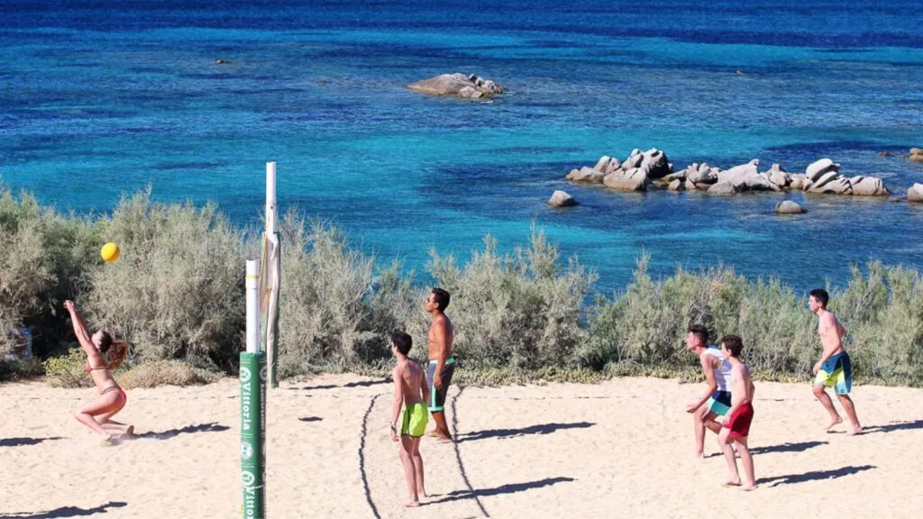 Gruppo di ragazzi che giocano a beach volley sulla spiaggia con il mare e le rocce sullo sfondo.