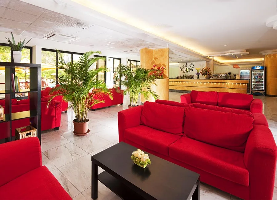 Hall dell'hotel con divani rossi, piante verdi e bancone bar sullo sfondo