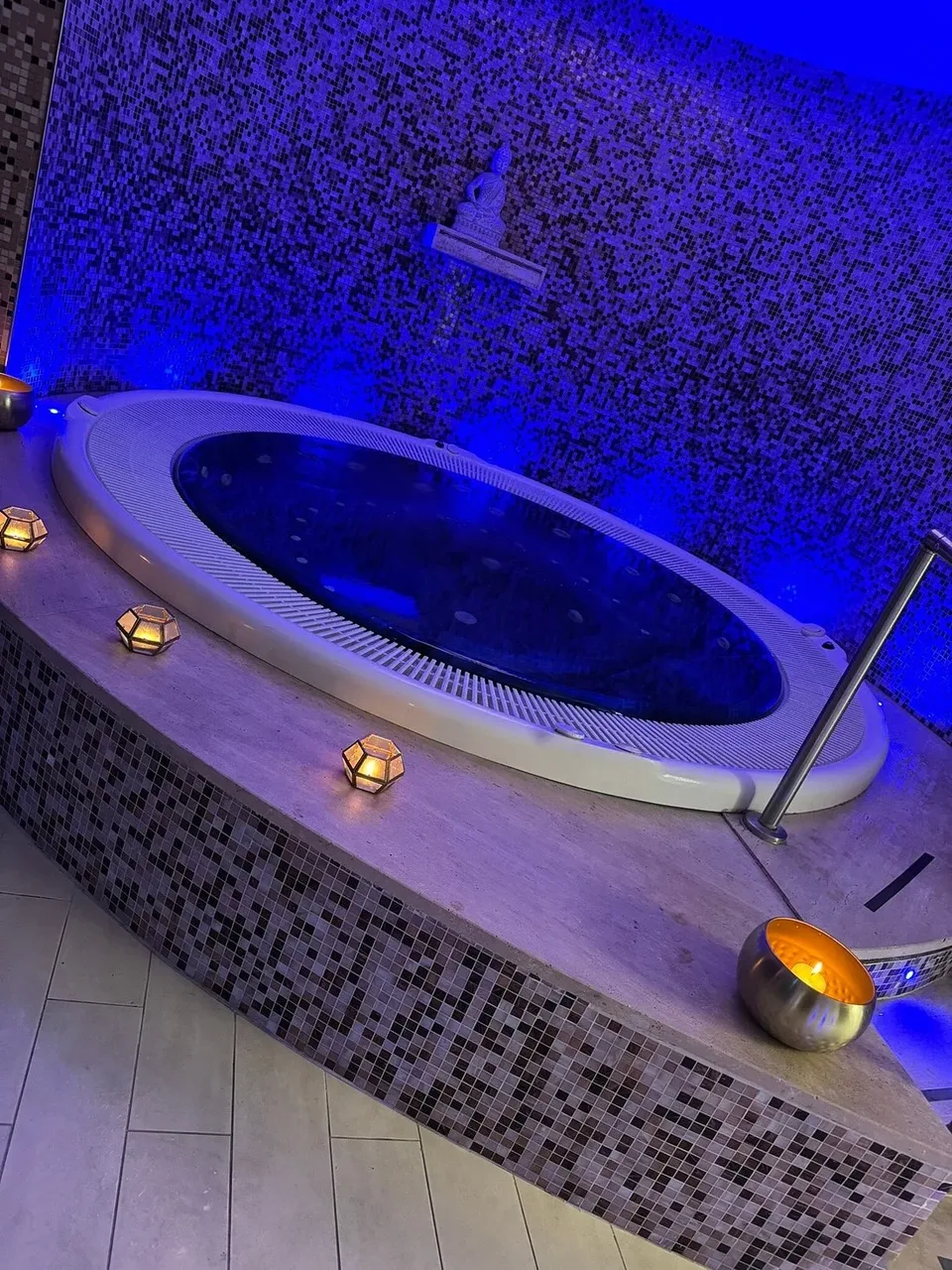 Vasca idromassaggio ovale con illuminazione blu e candele su parete a mosaico