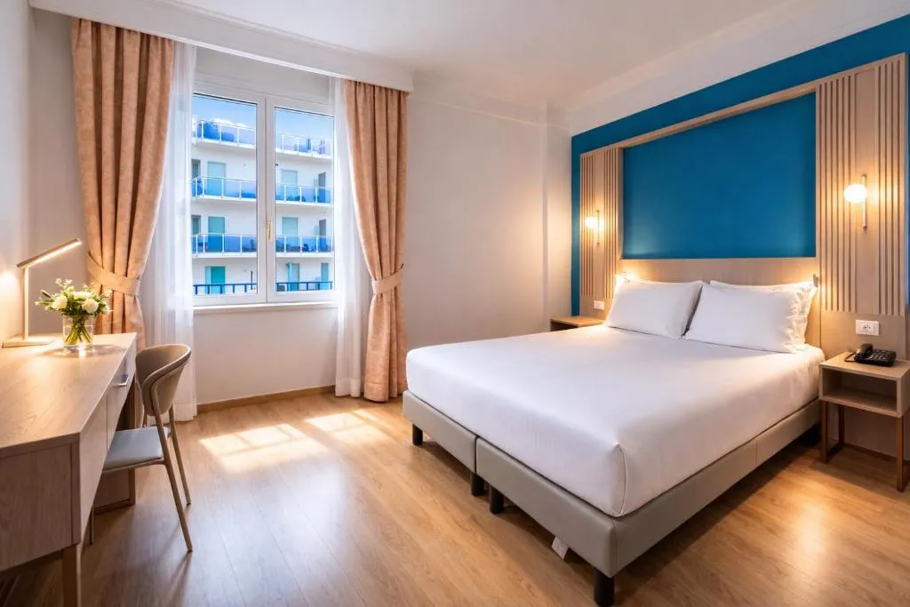 Camera moderna con testiera blu, scrivania, sedia e finestra con vista su edificio