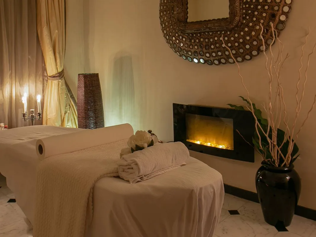 Sala massaggi spa con lettino, asciugamani arrotolati, camino decorativo e specchio etnico alla parete
