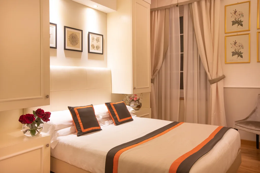 Camera accogliente con letto matrimoniale, cuscini arancioni, rose rosse e stampe botaniche
