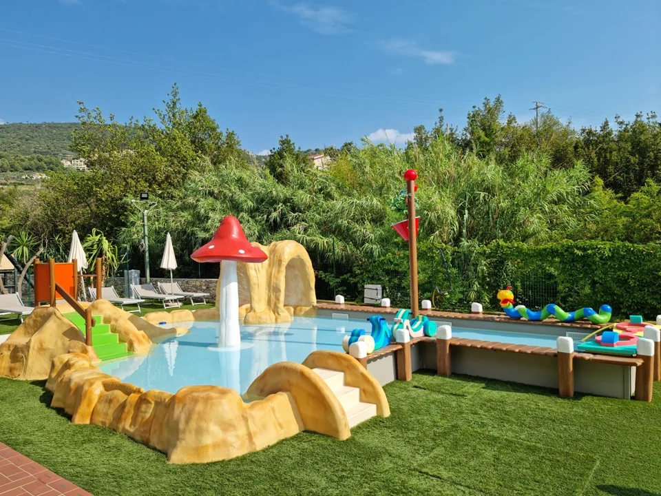 Piscina per bambini con fungo rosso e giochi acquatici circondata da vegetazione