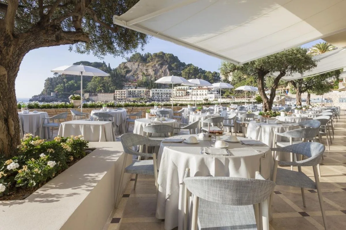 Ristorante all'aperto con tavoli bianchi sotto ulivi vista costa e mare