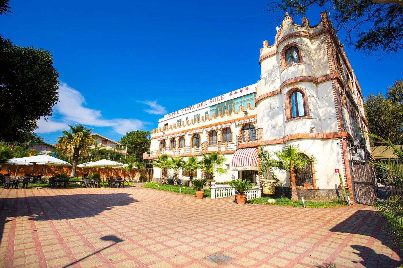 Facciata dell'Hotel Costa del Sole con torretta, palme e tavoli all'aperto