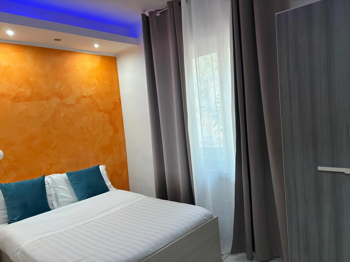 Camera matrimoniale con parete arancione, cuscini turchesi e illuminazione blu a soffitto